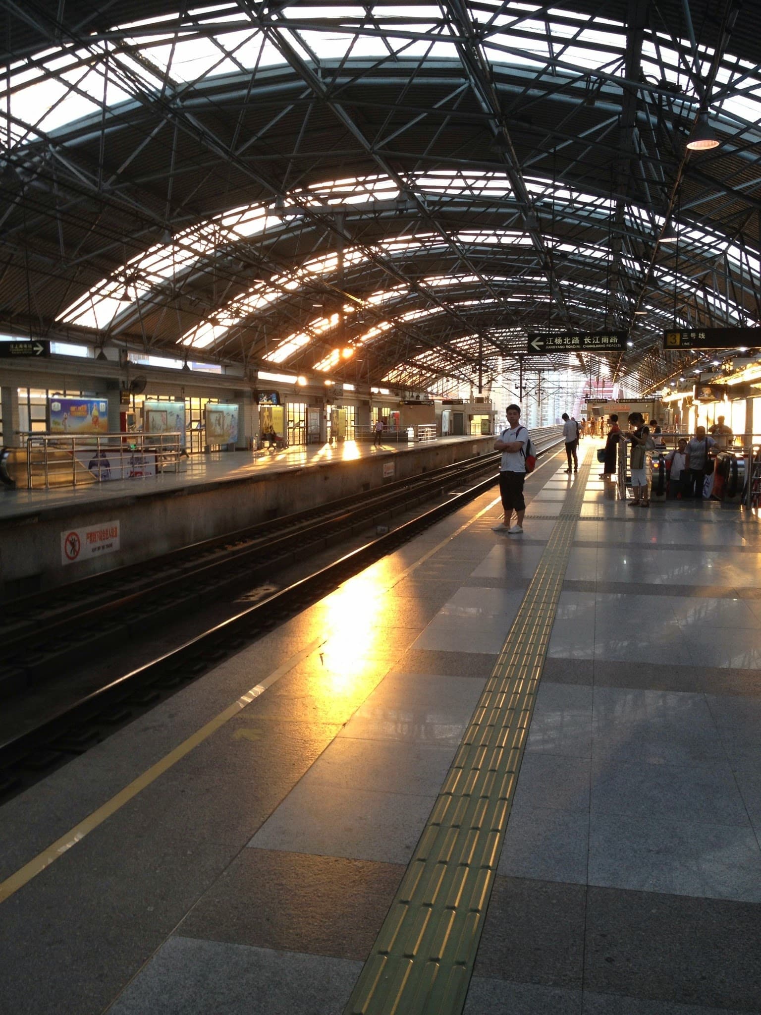 A bustling train station platform with a metal roof and a long train track, people waiting to board
