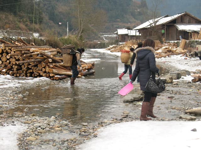People walking through a shallow stream in a snowy village with woodpiles and buildings
