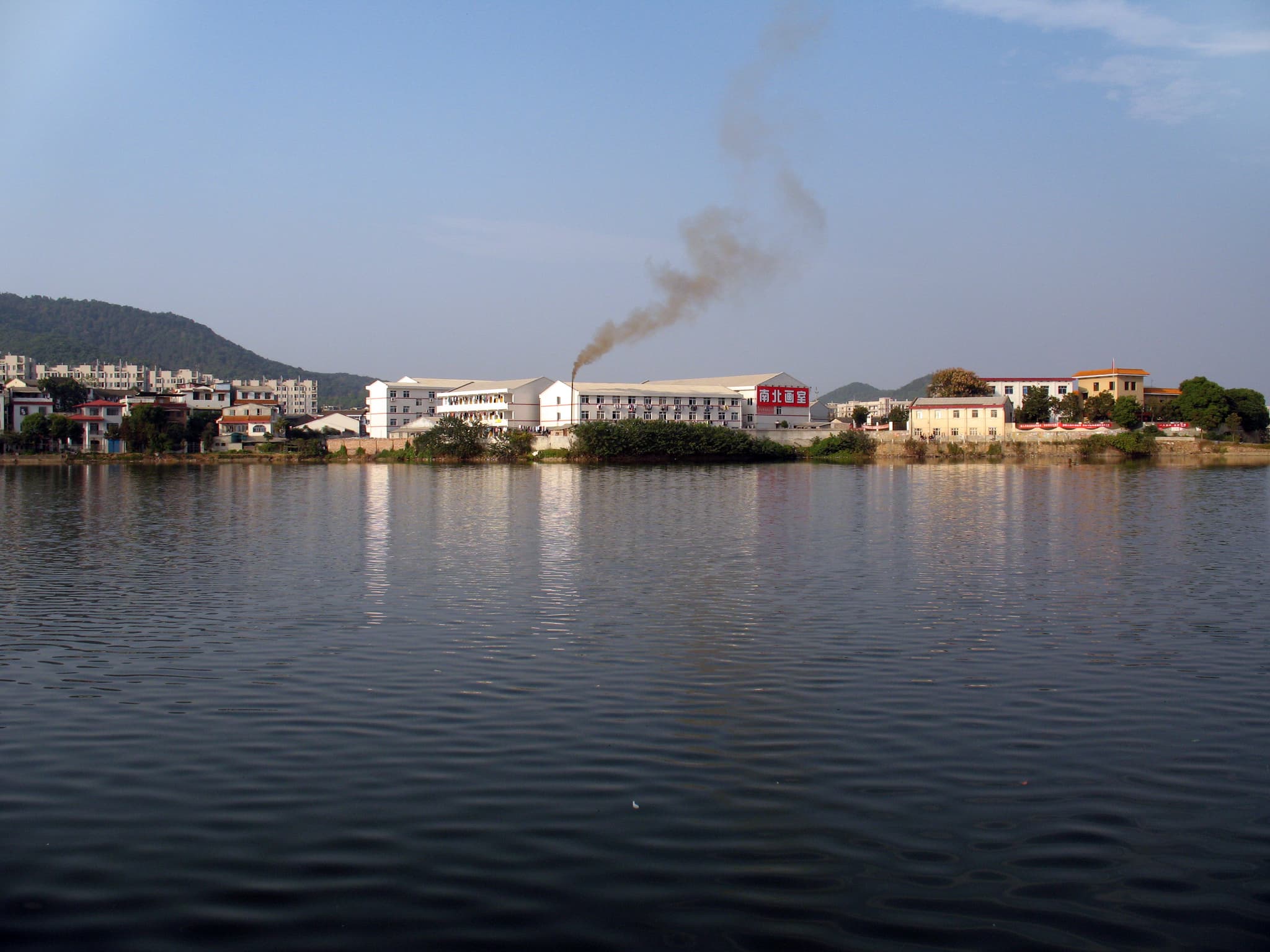 A train emitting smoke beside a serene lake with buildings and a hill in the background