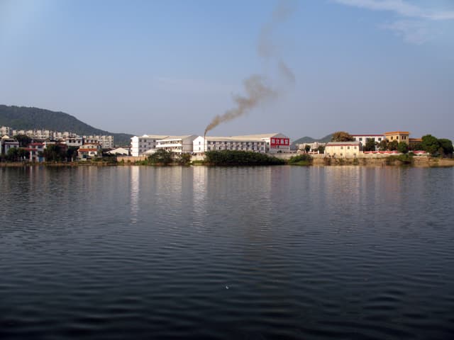 A train emitting smoke beside a serene lake with buildings and a hill in the background