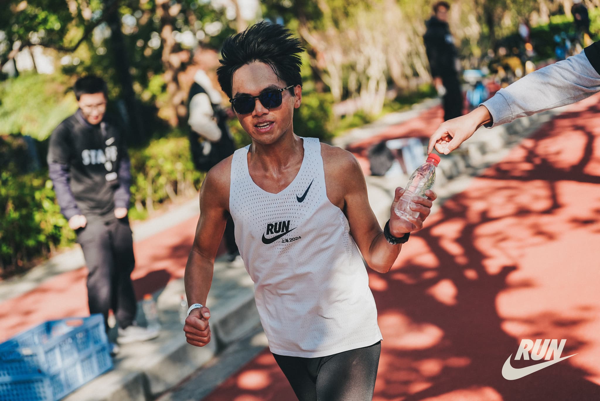 A man wearing a white Nike tank top and sunglasses runs on a track with other people in the background