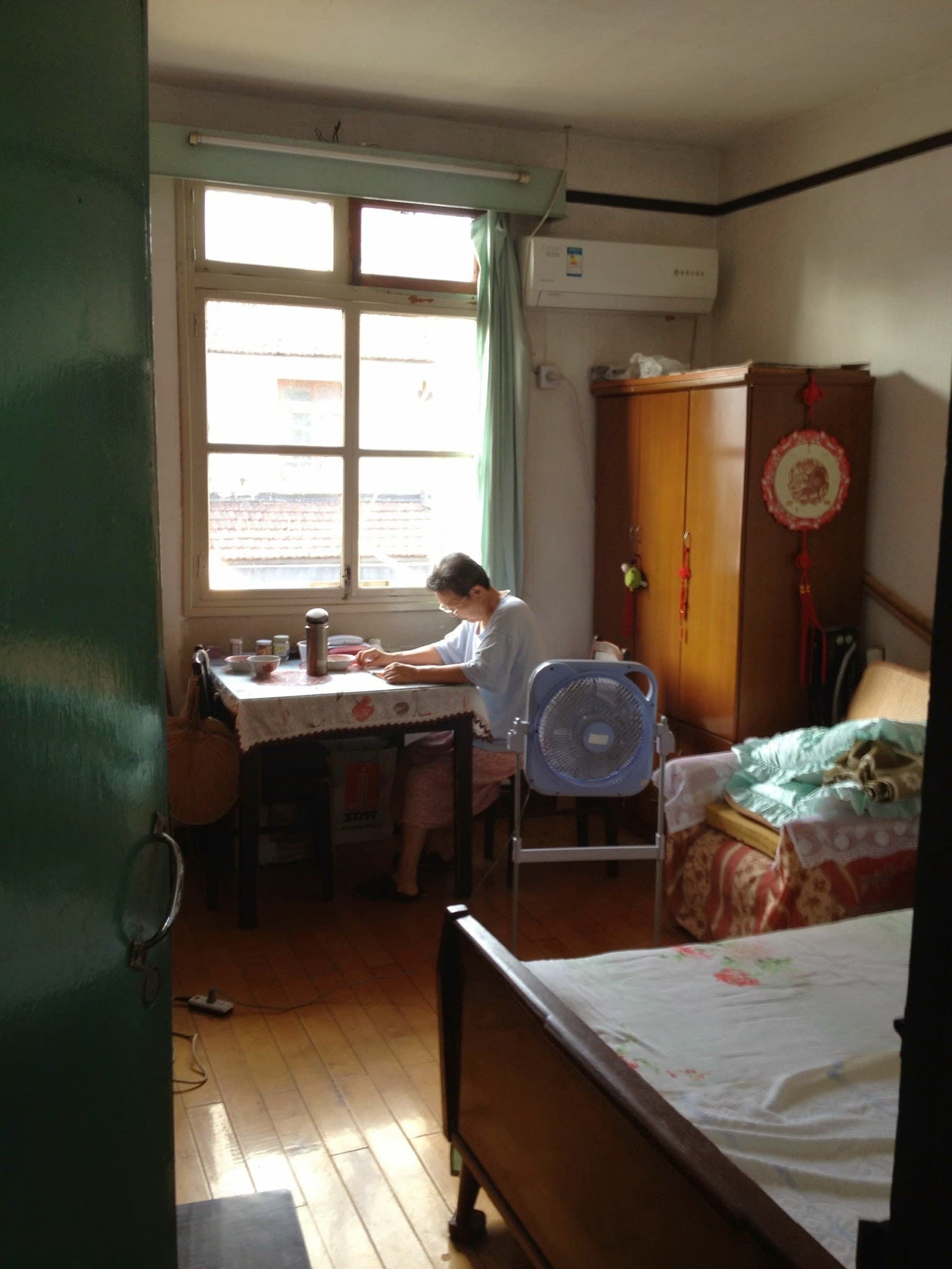 Person sitting at desk by window in small room with bed