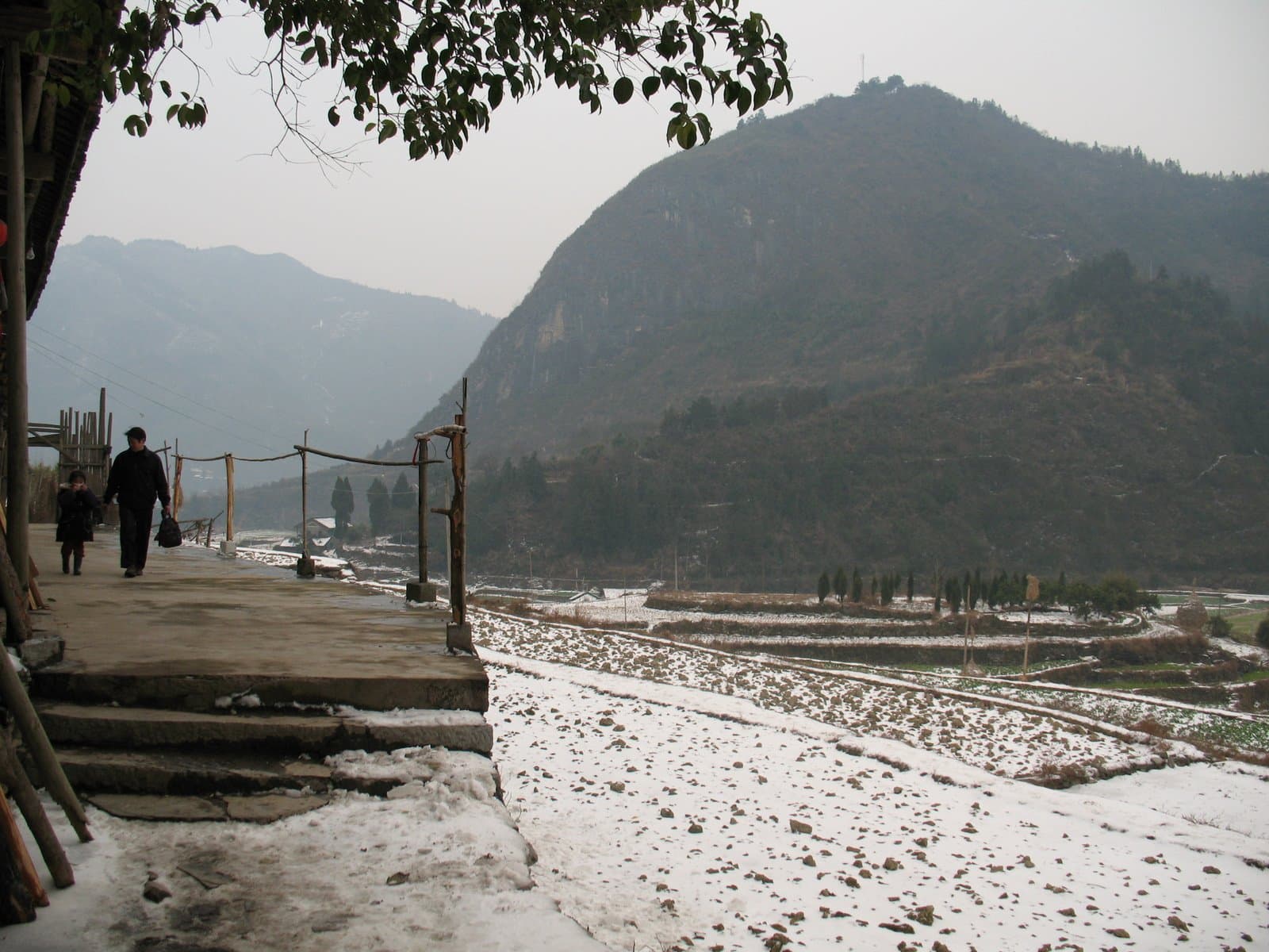 A snow-covered path leading to a mountainous landscape with a fence and tree in the foreground