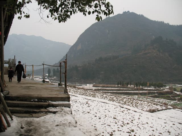 A snow-covered path leading to a mountainous landscape with a fence and tree in the foreground