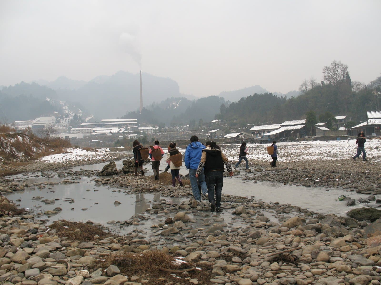 People walking through a rocky, muddy area with snow-covered mountains in the background on a foggy day