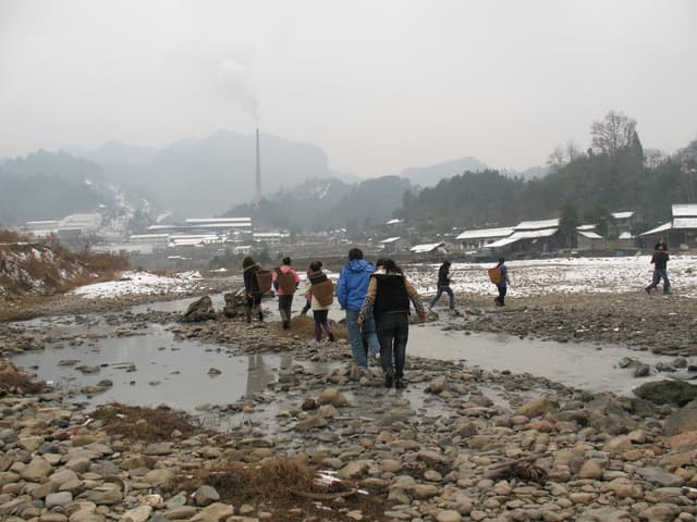 People walking through a rocky, muddy area with snow-covered mountains in the background on a foggy day