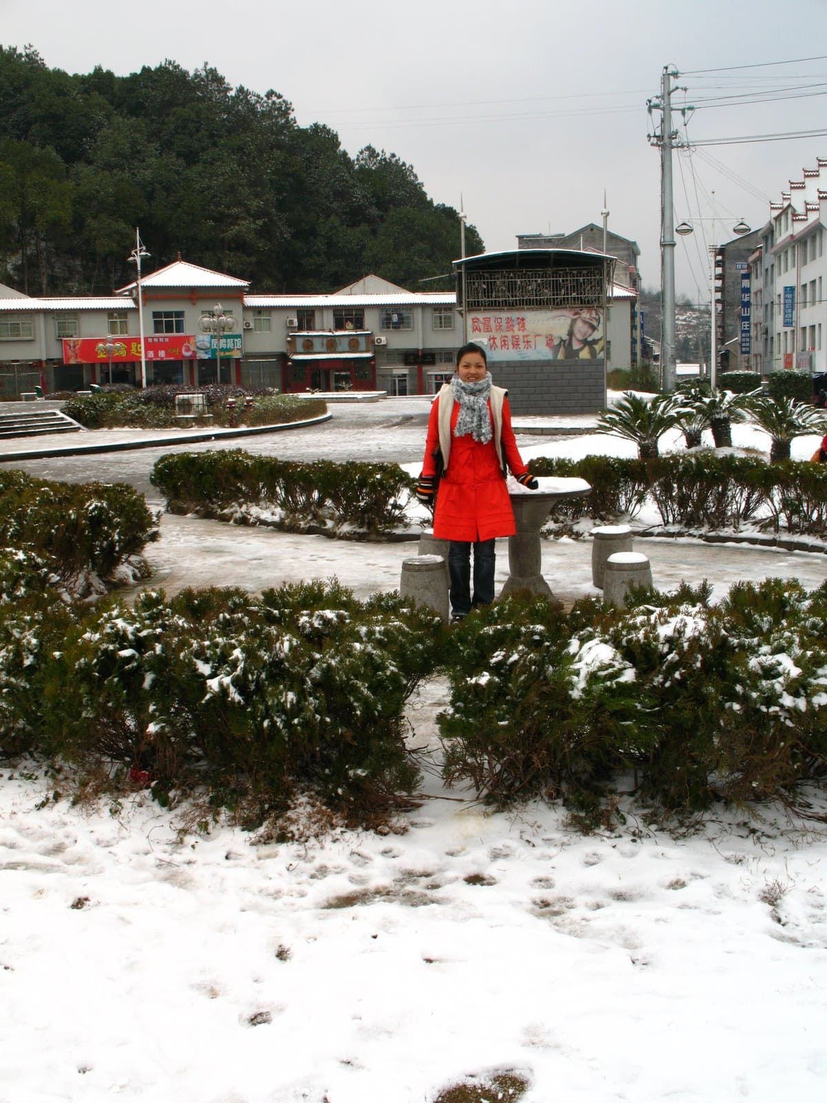 A woman in a red coat standing amidst snow-covered bushes and buildings