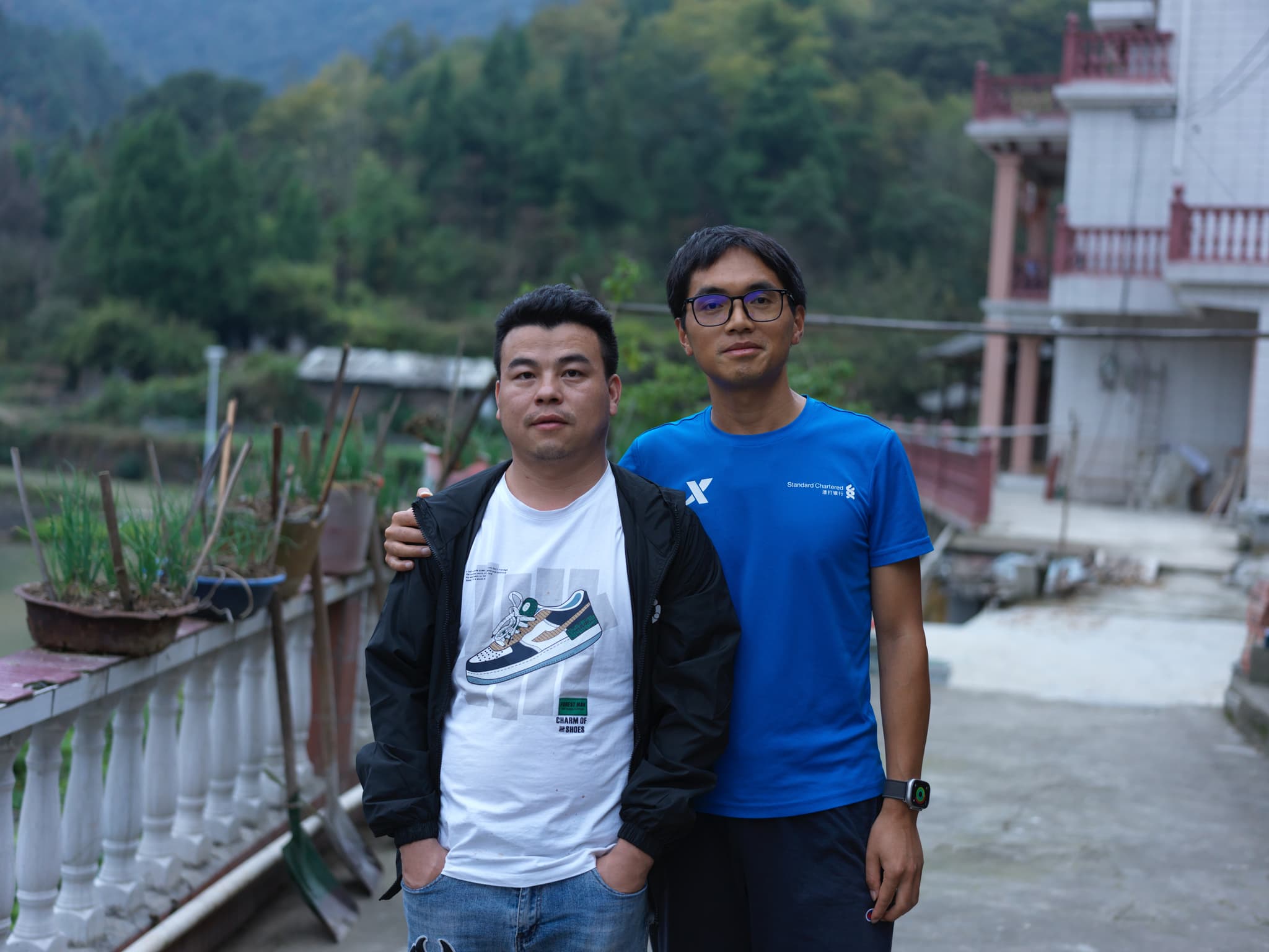 Two men standing together outdoors with a building and trees in the background
