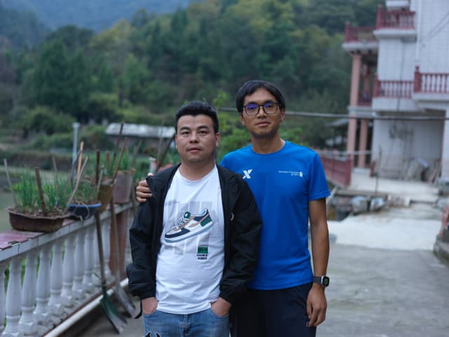Two men standing together outdoors with a building and trees in the background