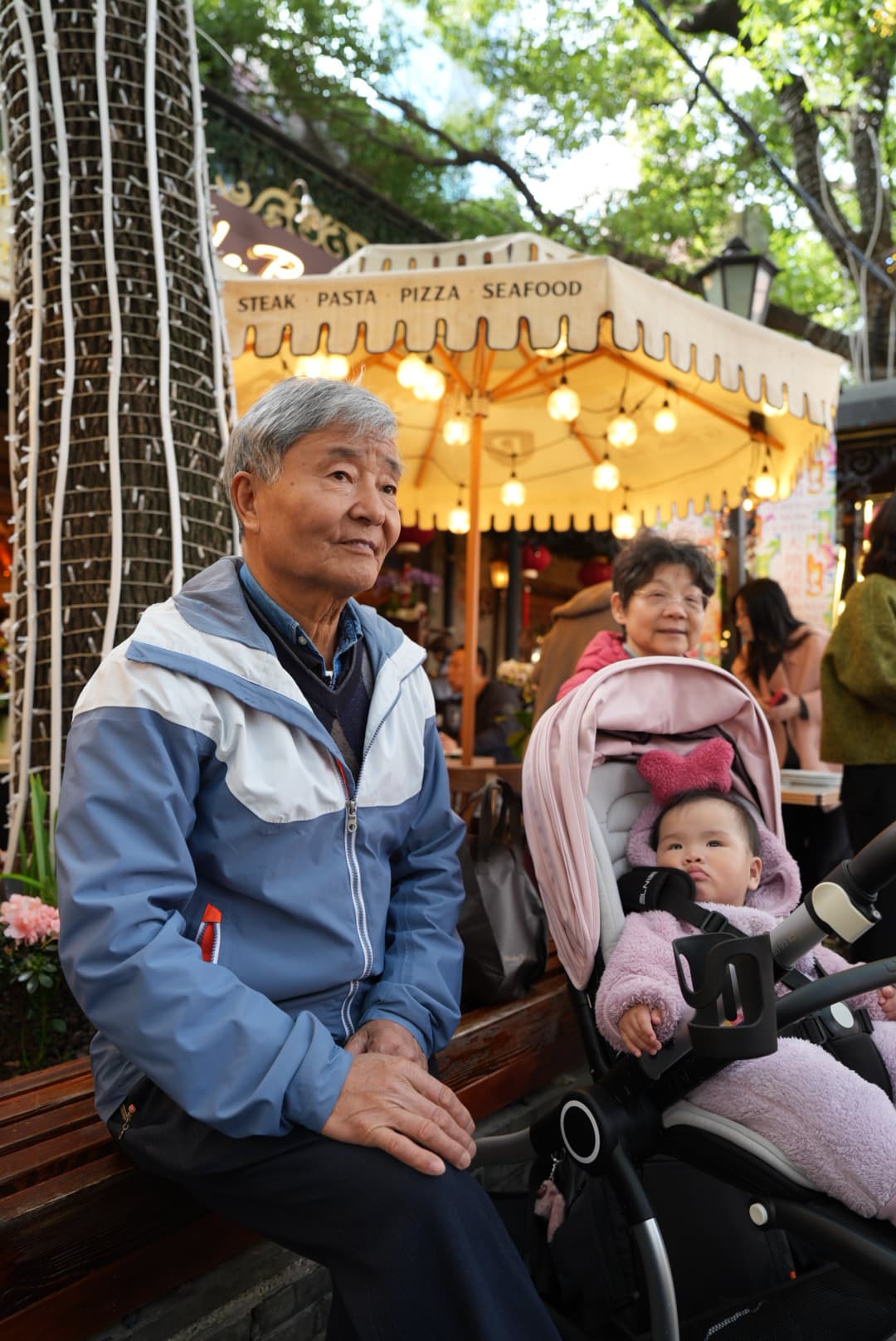 An older man pushing a stroller with a baby through a outdoor setting with trees and a pagoda-style building in the background