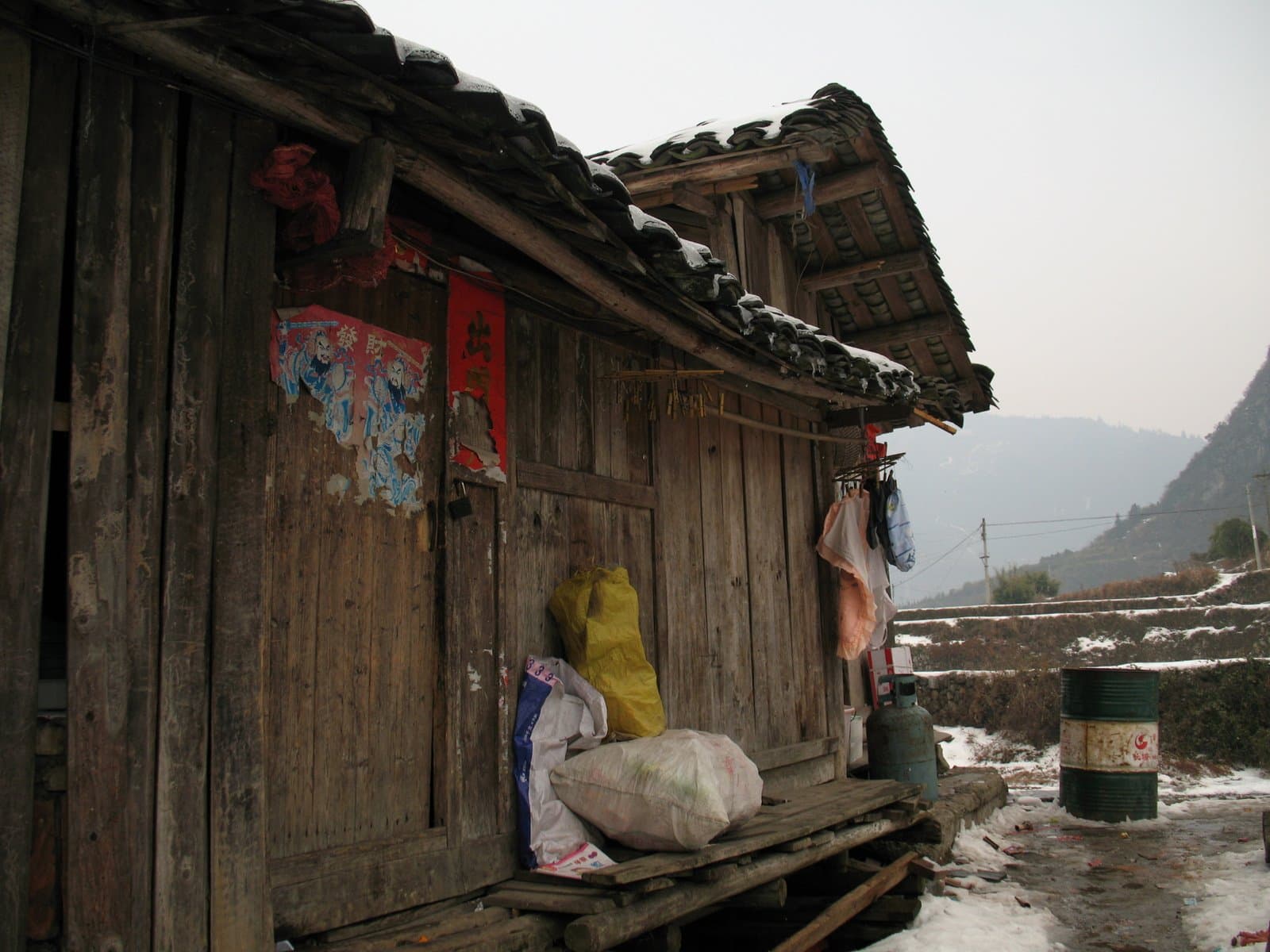 A rustic wooden hut with a tiled roof and weathered exterior, situated in a mountainous area with a barrel and bags nearby