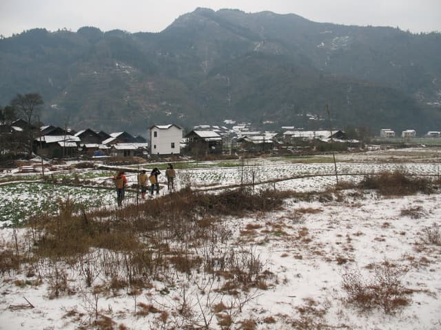A snowy field with people walking in the distance, a village and mountains in the background