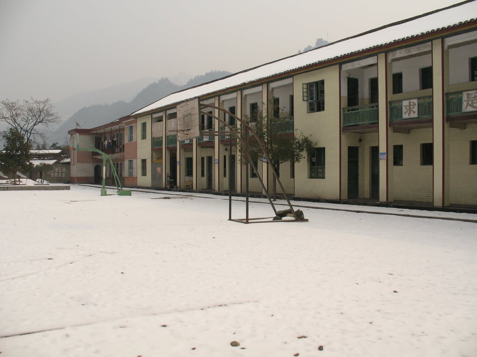 A long, two-story building with a snow-covered courtyard and mountains in the background on a foggy day