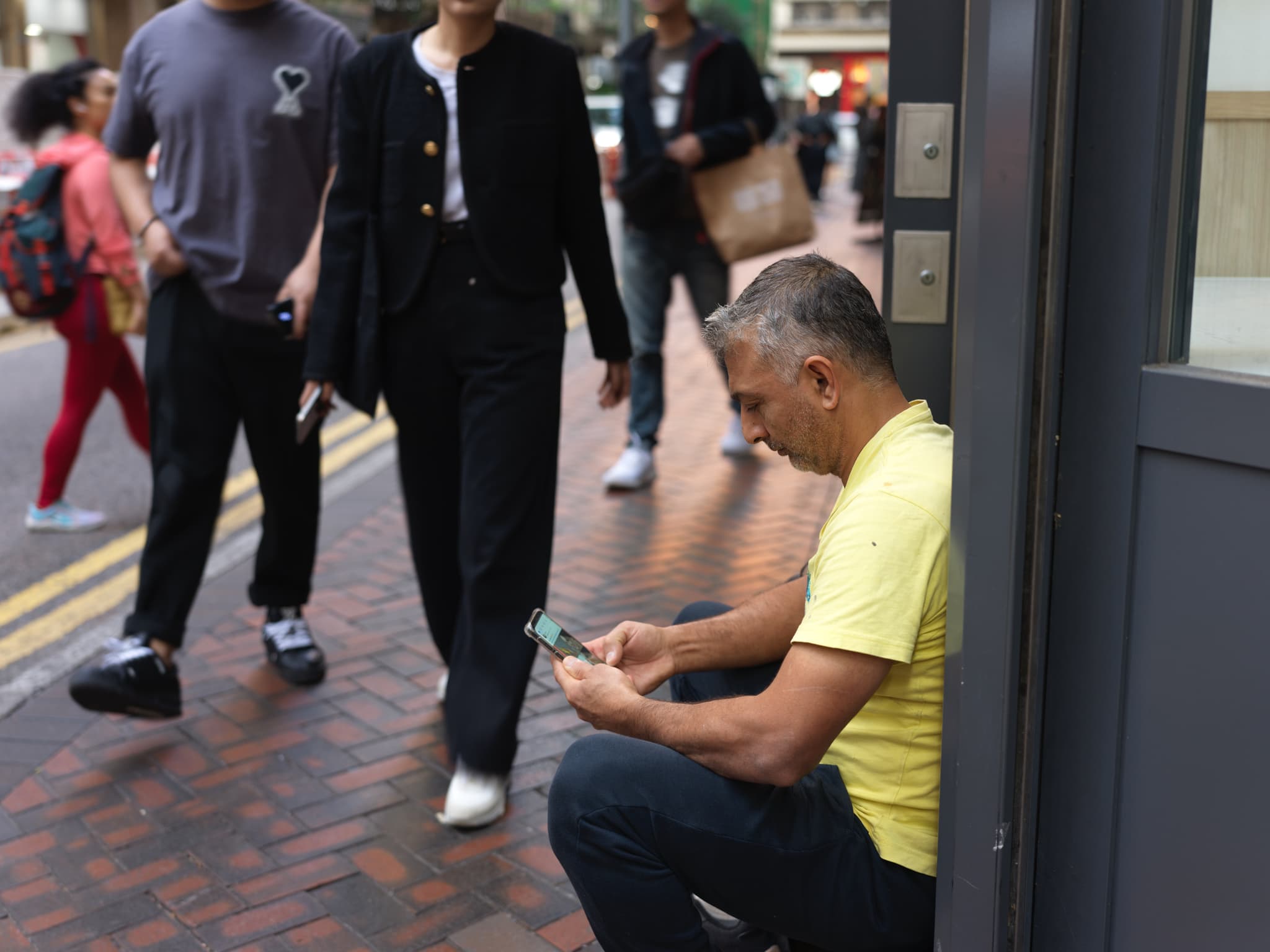man sitting on ledge looking at phone with people walking by on sidewalk
