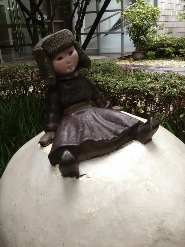 A vintage doll statue sits on a white sphere in a garden setting