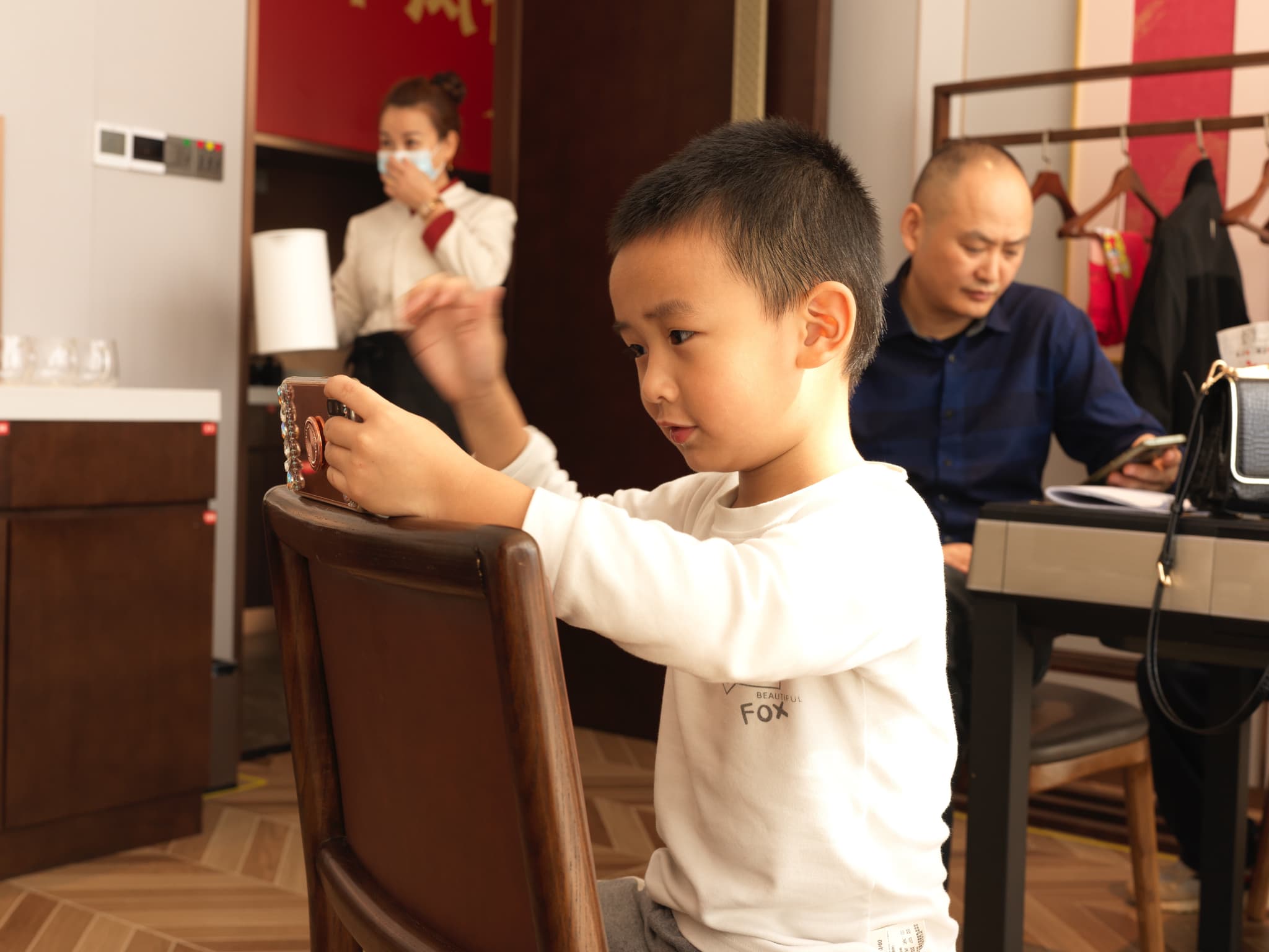 A young boy sitting on a chair, with a man and another person in the background